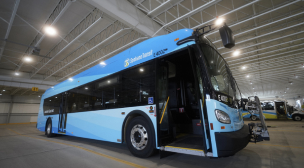 A light blue electric bus with the number E4002 is parked inside a spacious, well-lit transit facility. The bus door is open, revealing the interior. Branded with "Suburban Transit," it appears to be in a storage or maintenance area, adhering to the facility's style guide.