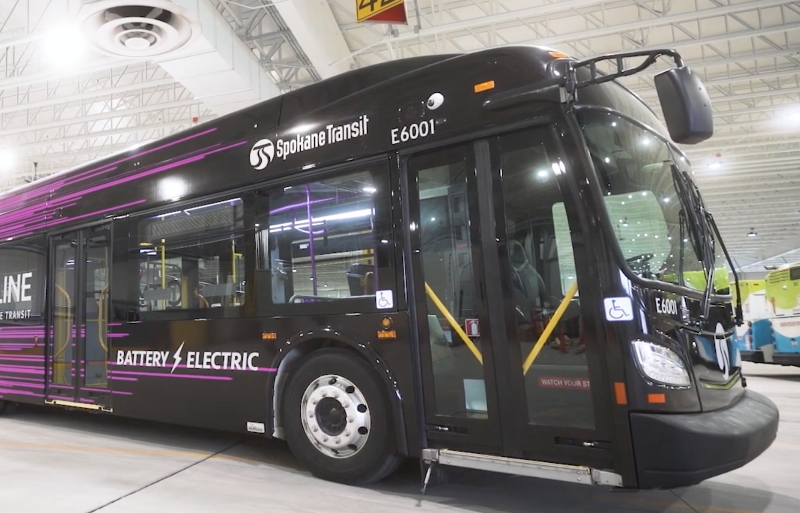 A modern battery-electric bus in a maintenance facility, featuring prominent Spokane Transit branding and a sleek black and purple design.