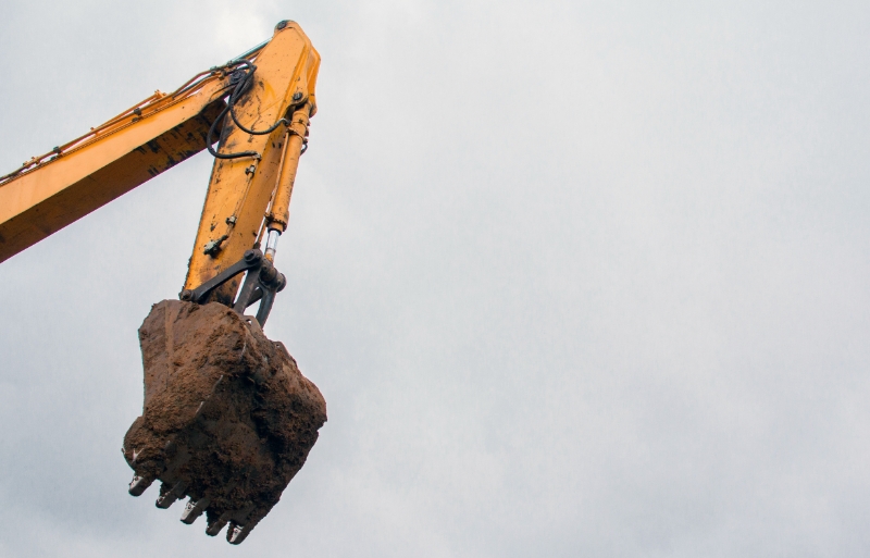 A yellow excavator arm with a metal claw holding a large clump of earth, set against a grey cloudy sky, near a Spokane Transit station.