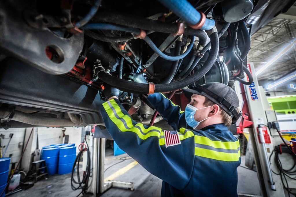 An STA mechanic wearing a mask and high-visibility clothing works on the underside of a vehicle in a garage, amidst various tools and equipment.