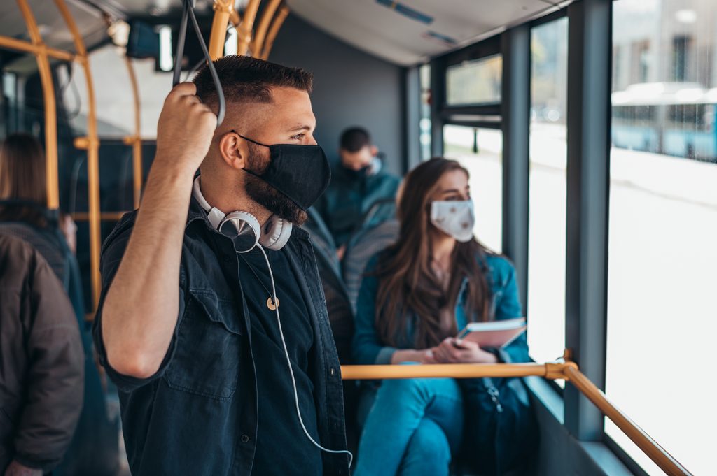 An image of a young man riding public transport, wearing a protective mask as he looks out the window.