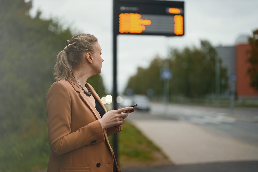An image of a woman standing outside by a bus stop, looking off into the distance as she waits for the bus to arrive