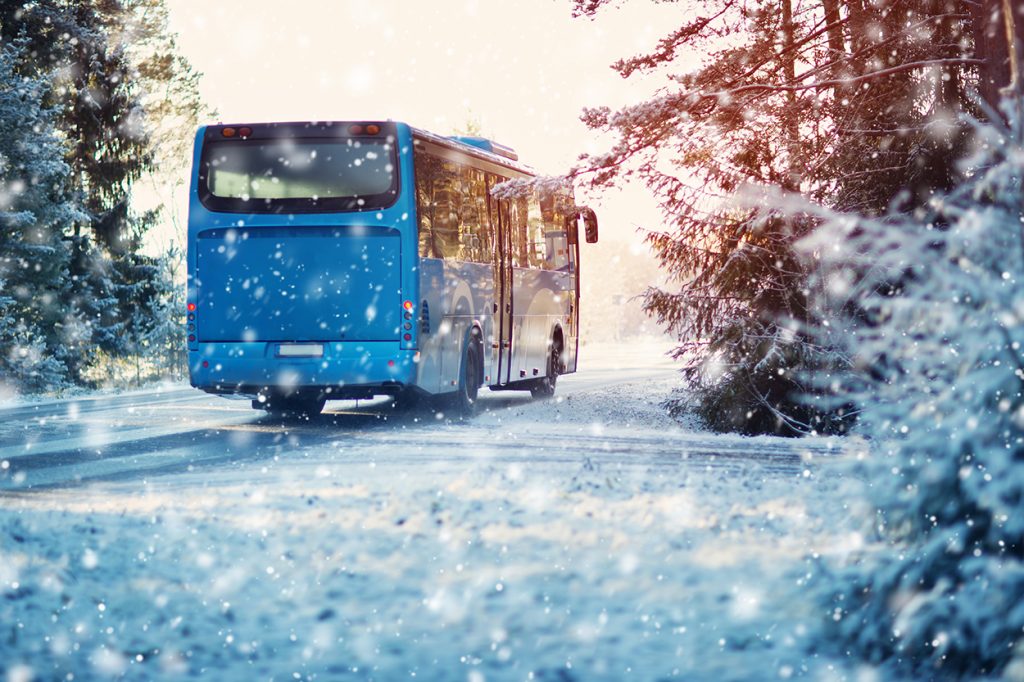 An image of a transit bus driving into the sunrise on a snowy road during winter.