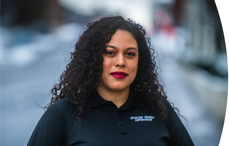 A confident woman with curly hair and red lipstick wearing a black shirt labeled "Spokane Transit Supervisor," stands outdoors with a blurred street background.