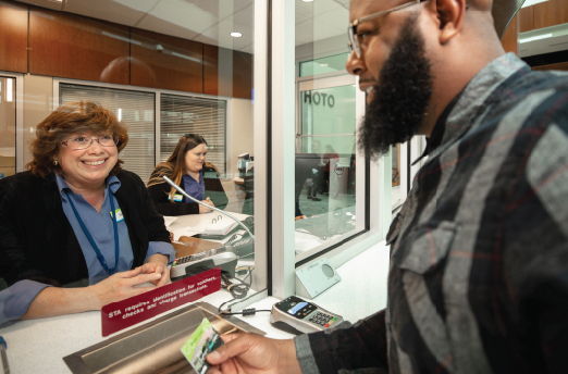 A cheerful female pharmacy assistant hands a man his credit card at a Spokane pharmacy counter, where another worker can be seen in the background.