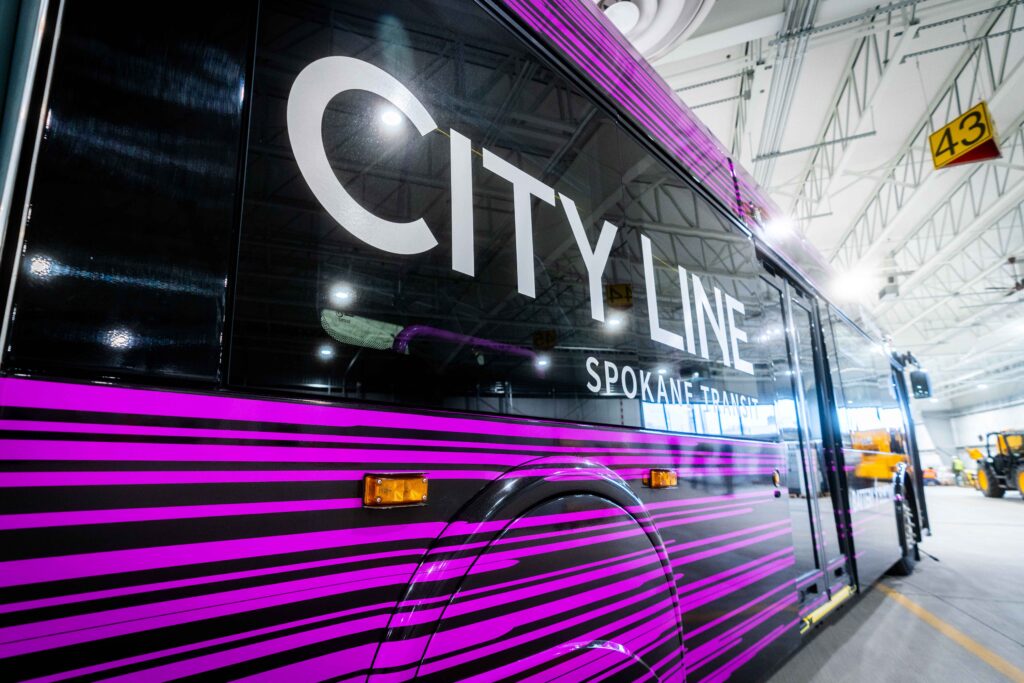 A vibrant pink and purple striped bus with "Spokane Transit" on the side, parked inside a maintenance facility with high ceilings and visible workshop areas.