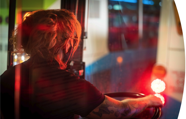 A Spokane Transit bus driver with tattoos and glasses is seen from behind, steering a bus at night, with blurred city lights and reflections visible through the window.