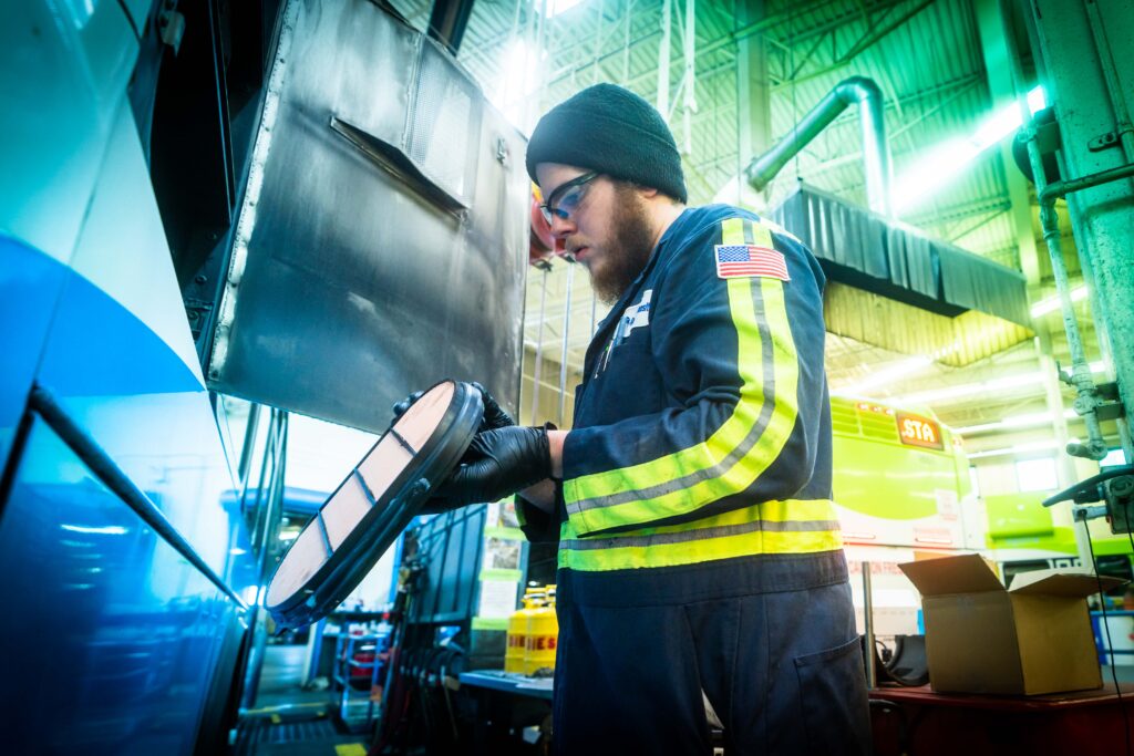 A worker in a reflective vest and beanie inspects machinery in an industrial setting, with vibrant lighting and large equipment surrounding him, including Spokane Transit vehicles.