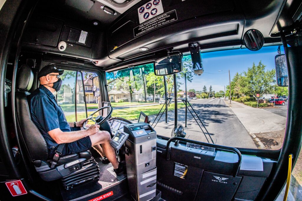 A Spokane Transit bus driver is seated at the wheel of a bus, navigating a suburban street. The dashboard and passenger area are visible, with a wide windshield providing a clear view of the road and houses