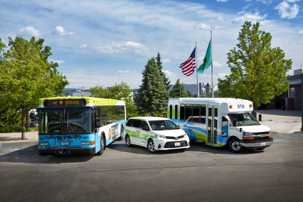 an STA Bus, Vanpool Van, and Paratransit Van in front of the flags of the United States and Washington State.