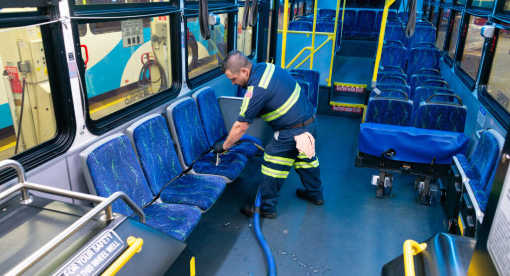 A worker in a reflective vest uses a cleaning hose to wash the floor and seats of an empty Spokane Transit bus, showing a careful cleaning process.
