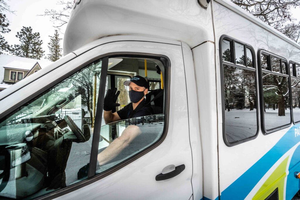 A Spokane Transit bus driver, wearing a mask and gloves, waves from the driver's seat of a bus on a snowy day, surrounded by winter trees.