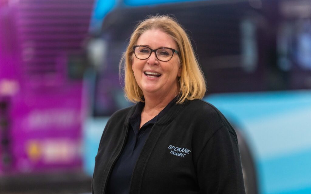 A cheerful middle-aged woman with shoulder-length blonde hair, wearing glasses and a Spokane Transit uniform, smiling in front of colorful buses.
