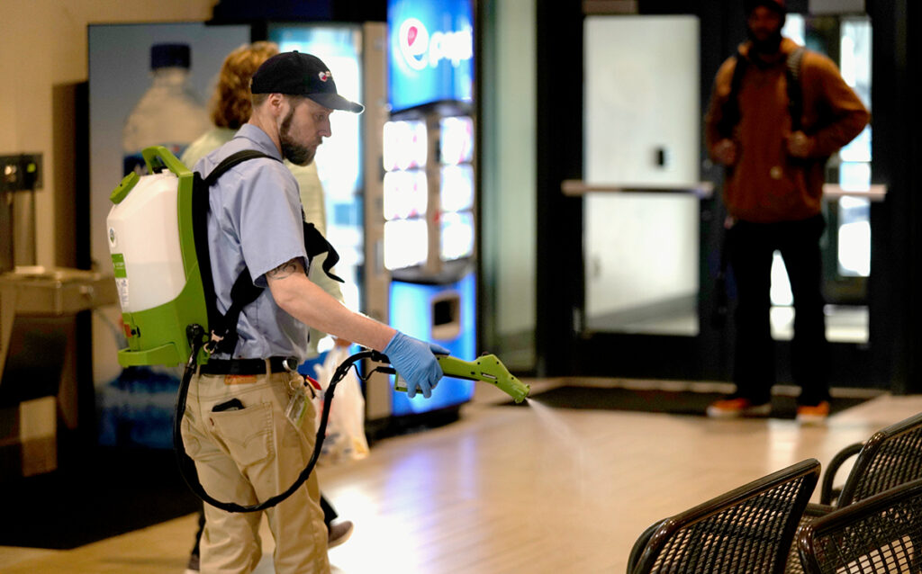 A man wearing a cap and a backpack sprayer disinfects a public area inside a Spokane Transit building, near a seating area and a digital screen, while another man observes from a distance.