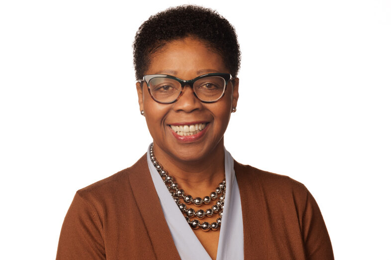 A woman with short curly hair, wearing glasses, a gray necklace, and a brown blazer over a light blue top, smiles warmly against a plain white background, exuding the confidence of someone ready to join the board of directors.