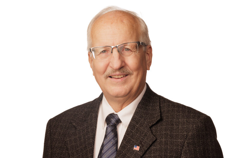 A smiling elderly caucasian man wearing a dark blazer, white shirt, blue tie, and glasses against a plain white background. He has a mustache and wears a Spokane Transit flag pin on