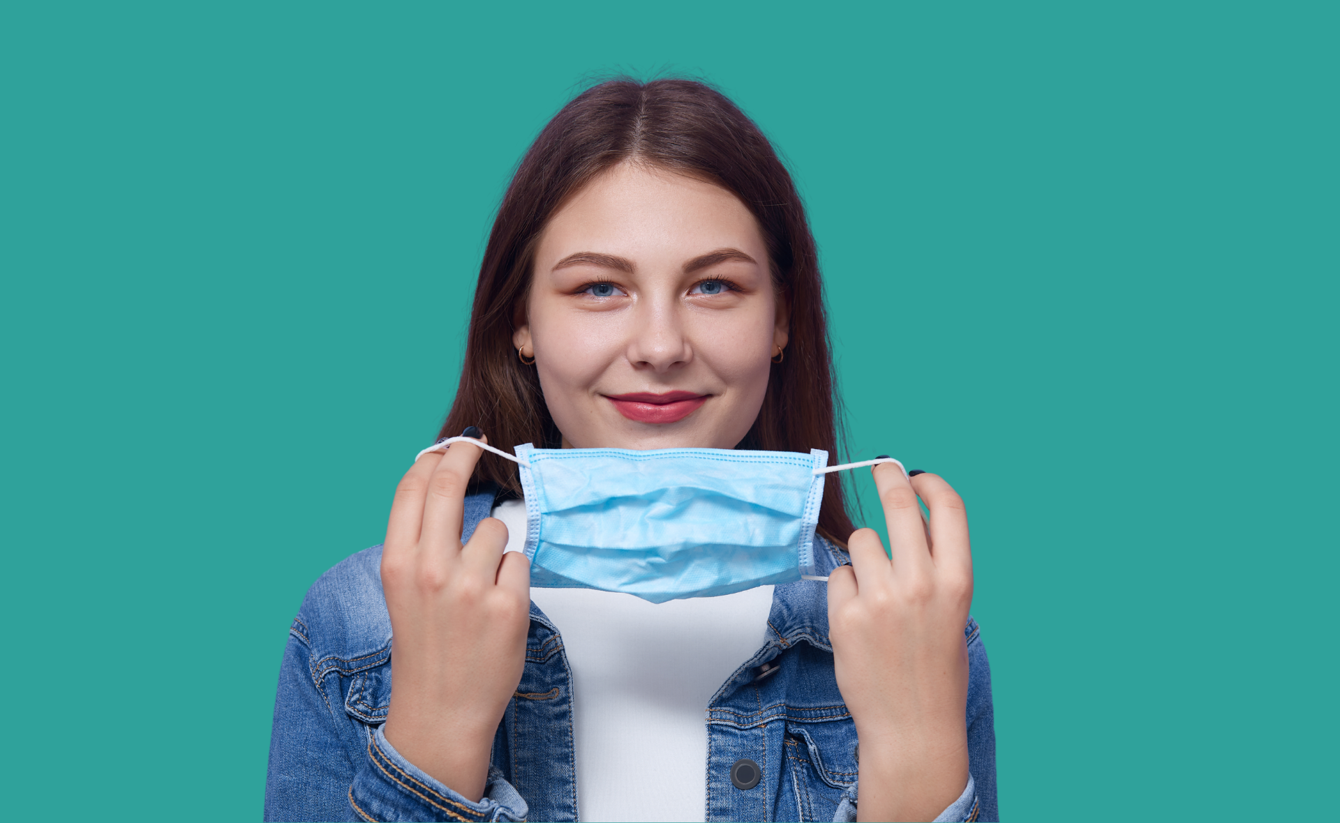 A young woman holding a face mask, smiling and looking at the camera. She wears a denim jacket, isolated on a turquoise background, promoting Spokane Transit's health safety initiatives.