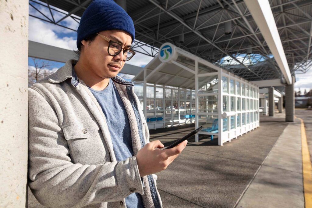 A person wearing a blue beanie, glasses, and a gray jacket leans against a concrete pillar while looking at a smartphone. They stand near a modern bus stop with clear panels and a blue sign overhead under a large metal roof structure.