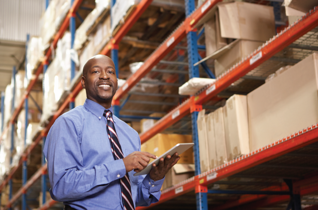 A smiling black man in business attire holding a tablet in a Spokane transit warehouse, with shelves stocked with boxes in the background.