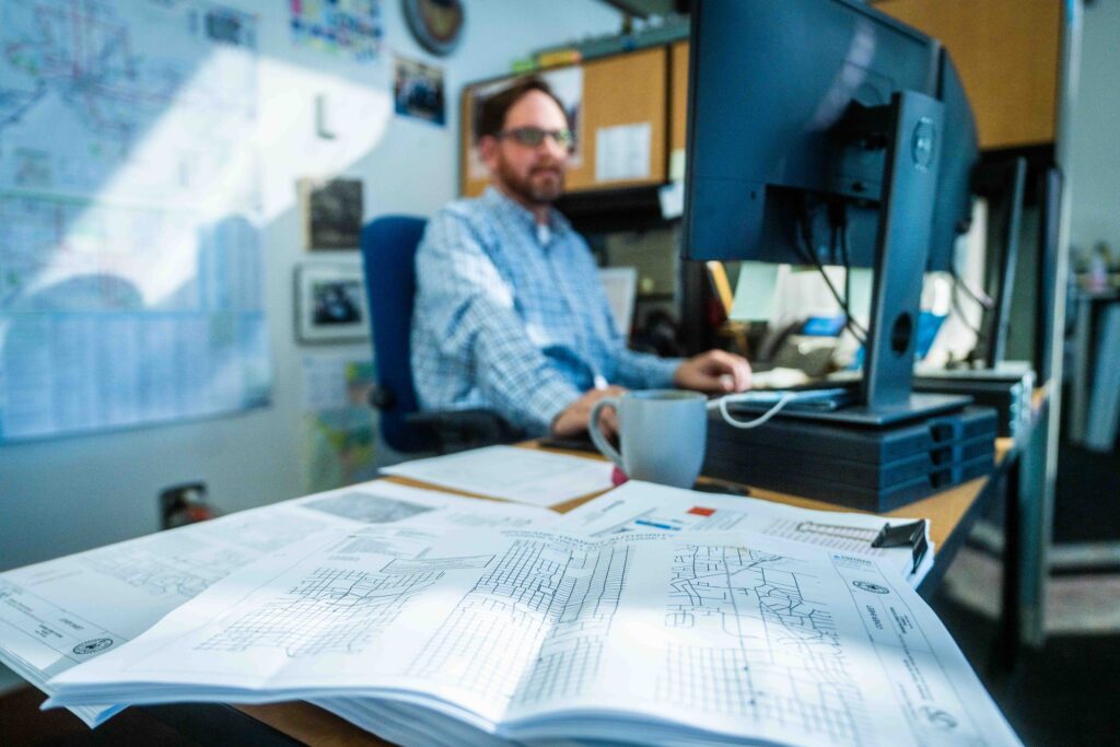 Plans on Desk with Man at computer behind.