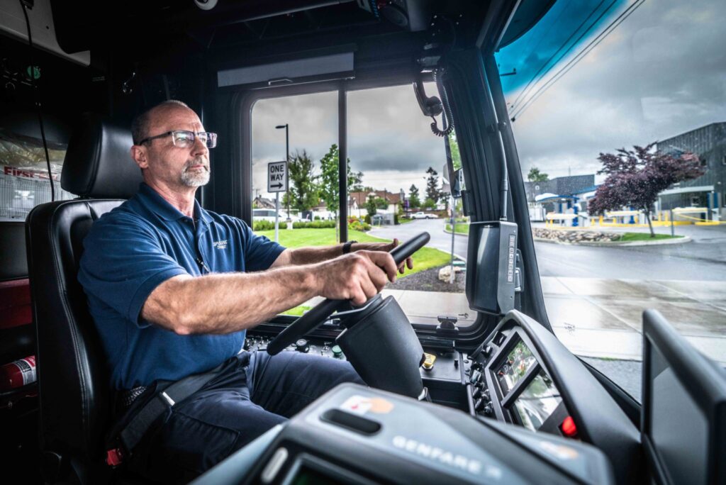A bus driver wearing a blue shirt and glasses focuses on the road while driving. The bus interior is visible with a steering wheel, dashboard, and controls. Outside the window, a rainy day scene with a "one way" sign, trees, and buildings in the background.