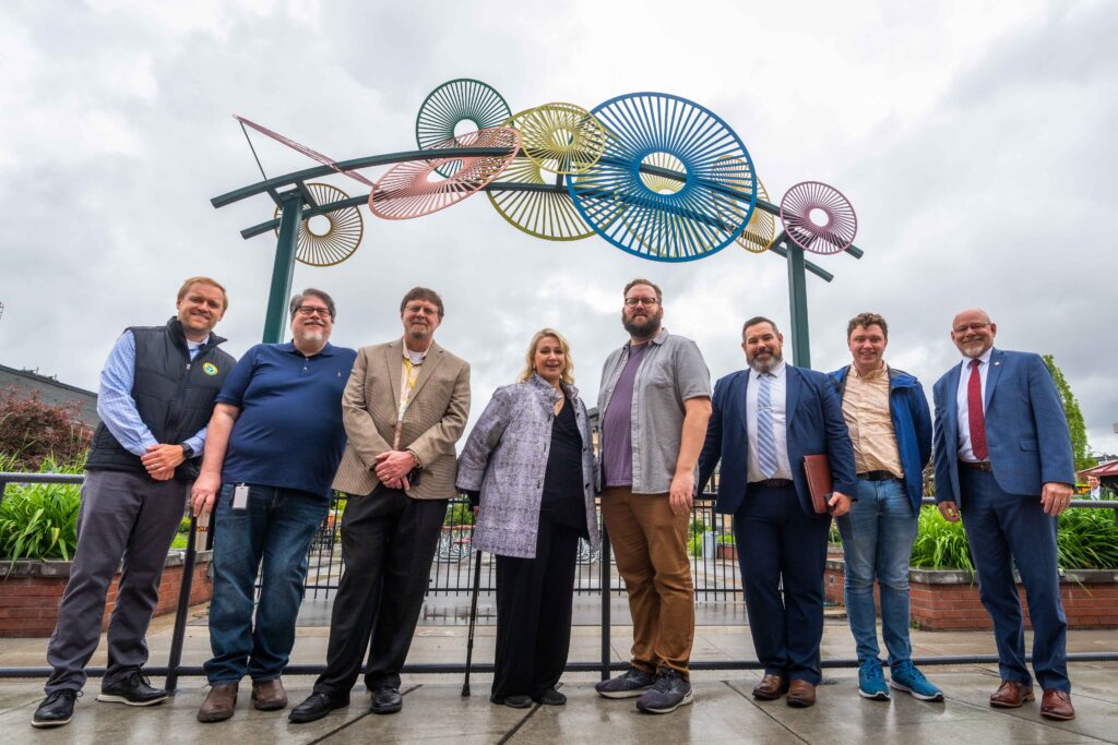 A group of eight people stands smiling under an outdoor sculpture made of circular and semi-circular materials in various colors. The sky is overcast, and they are dressed in business casual attire. The background features some greenery and a brick structure.