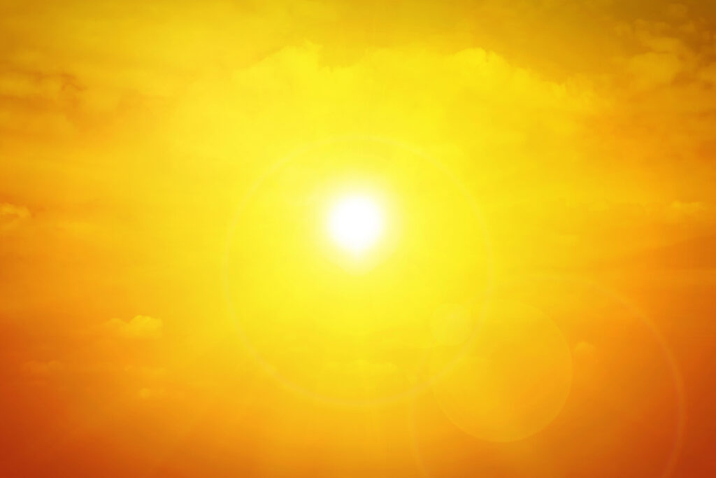 Bright yellow sun glowing intensely in a clear sky with a warm golden hue, diffusing light across the image. Few faint clouds are barely visible around the sun, enhancing the overall warmth and radiance. Sun flares create lens effects in the foreground.