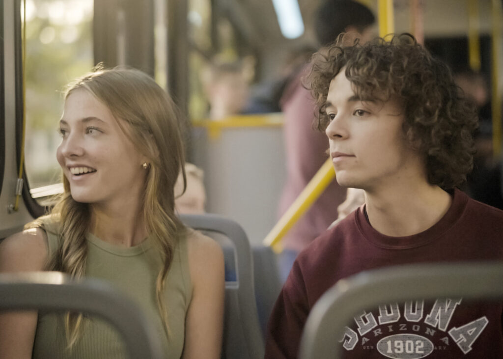 A young woman with long blonde hair wearing a green sleeveless top, and a young man with curly brown hair wearing a maroon sweatshirt sitting together on a bus. The woman is smiling and looking out the window while the man looks straight ahead.