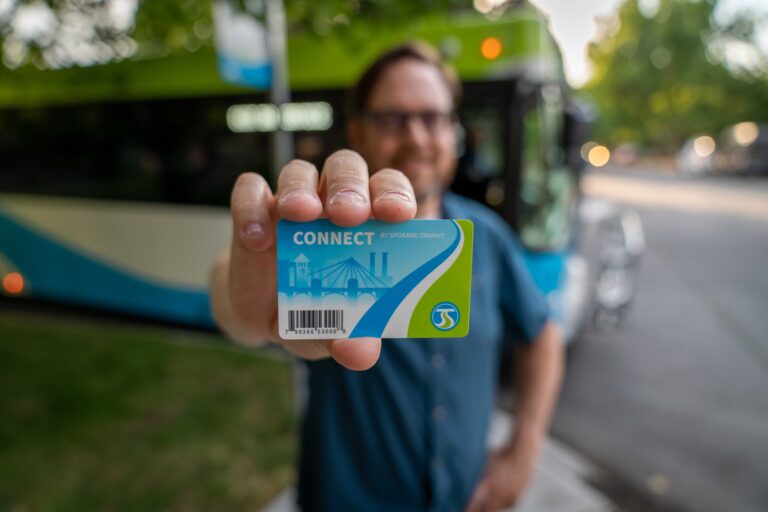 A person standing near a green and blue bus holds a Spokane Transit "Connect" card towards the camera. The person is slightly out of focus, while the card is prominent and sharp, displaying a barcode, cityscape icons, and the Spokane Transit logo.