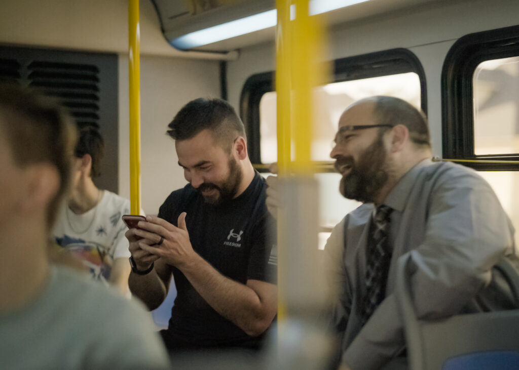 Two men are sitting on a bus, smiling and engaged in a lighthearted moment. The man on the left is looking at a smartphone and laughing, while the man on the right, wearing glasses and a tie, also smiles warmly. The bus's interior and yellow poles are visible.