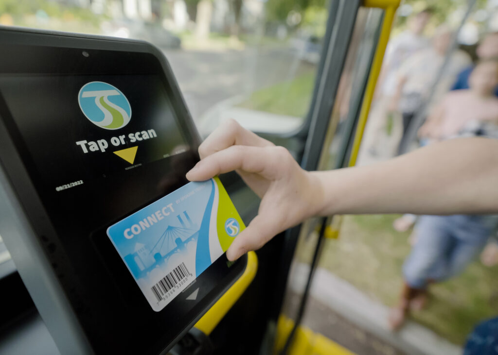 A person taps a blue Spokane Transit card on a black card reader labeled with a "tap or scan" sticker inside a bus, with passengers visible in the background.