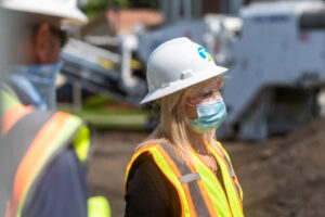 A woman in a construction site is wearing a white hard hat, a face mask, and a neon orange safety vest. She is accompanied by another person in similar attire, both diligently working on the new City Line project. Machinery and construction materials are visible in the background.