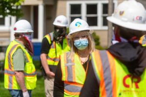 A group of construction workers wearing neon safety vests, hard hats, and face masks are gathered outdoors along the City Line. A person in the middle with a white hard hat and glasses appears to be speaking to others. A house with beige walls and white-trimmed windows is in the background.