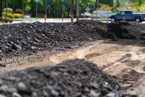 A construction site with piles of black asphalt chunks on a partly compacted dirt road sits near the city line. In the background, a turquoise pickup truck is parked near some traffic cones and signs, all under a canopy of green trees. The scene is brightly lit by sunlight.