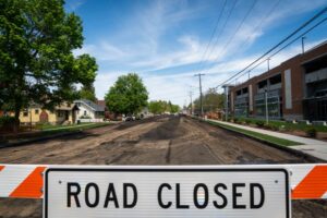 A road is under construction, with a "Road Closed" sign in the foreground. The street is dug up, and no vehicles are present. Trees and buildings line the city line under a clear blue sky.