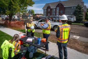 Construction crew members wearing safety vests and helmets working at a construction site near the City Line residential area. One person sits at a table with equipment, while others stand and discuss. A parked truck and construction machinery are visible in the background.