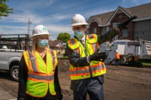 Two people, wearing white hard hats, face masks, high-visibility vests, and black gloves, stand at a construction site along the City Line. The person on the right gestures and points as equipment and workers operate in the background.