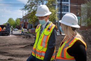 Two construction workers wearing safety vests, helmets, and face masks converse at a construction site. They stand on a partially paved road along the bustling city line, with buildings, trees, and equipment in the background.