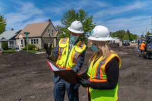 Two construction workers in safety gear and masks examine documents at a residential construction site near the City Line. The man gestures with one hand while holding a small tablet, and the woman holds a clipboard. Houses and construction vehicles are visible in the background.