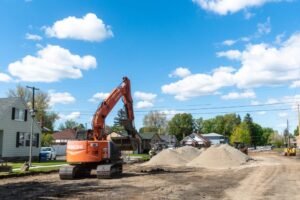 An excavator operates at a suburban construction site on a sunny day, surrounded by piles of dirt and gravel. In the background, the city line blends with houses and trees, while the sky above is dotted with scattered clouds.