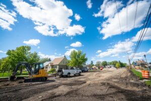 A road construction site with heavy machinery, including an excavator and a utility truck, sits along the City Line. A partially paved road runs alongside houses with green lawns and trees under a blue sky with clouds. Orange and white construction barrels line the road.