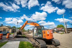 A bright orange excavator labeled with "Hitachi" and "CR" operates on a residential construction site with dirt and gravel piles. There are orange safety cones nearby, power lines above, and houses and trees under a partly cloudy blue sky, marking the city line in the distance.