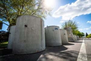 Large concrete cylinders are lined up on the side of a sunny road, forming part of the city line. The street is mostly empty, and trees provide a lush backdrop. The sky is blue with some clouds, and the sun shines brightly, casting shadows from the structures and trees.