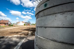 A large concrete cylinder in the foreground with a construction site in the background. The site features dirt and construction equipment. Residential houses line the city streets under a blue sky with scattered white clouds, forming a picturesque city line.