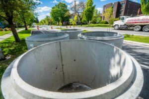 Concrete pipe sections are arranged on a closed street, with traffic cones visible in the background. A truck labeled "Cameron Reilly" is parked nearby. Trees and residential buildings line the city street. The sky is blue with some clouds, enhancing the tranquil city line.