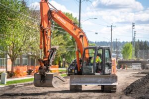 An orange Hitachi excavator operated by a worker in a reflective vest works on a construction site near the city line on a sunny day. The site is adjacent to a street with power lines, trees, and orange safety fencing.