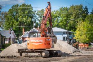 An orange Hitachi excavator is parked on a dirt road at a construction site, with its arm raised above a mound of sand. In the background, houses, trees, and construction barriers are visible under a partly cloudy sky and the distant city line.