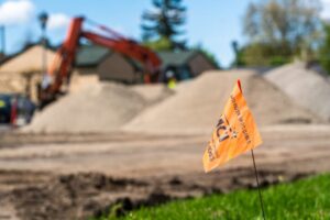 An orange flag labeled "WCI" marks a construction zone with sand piles and an excavator in the background. The city line is visible beyond, with trees and buildings in the distance under a clear sky with a few clouds. The foreground features a patch of green grass.