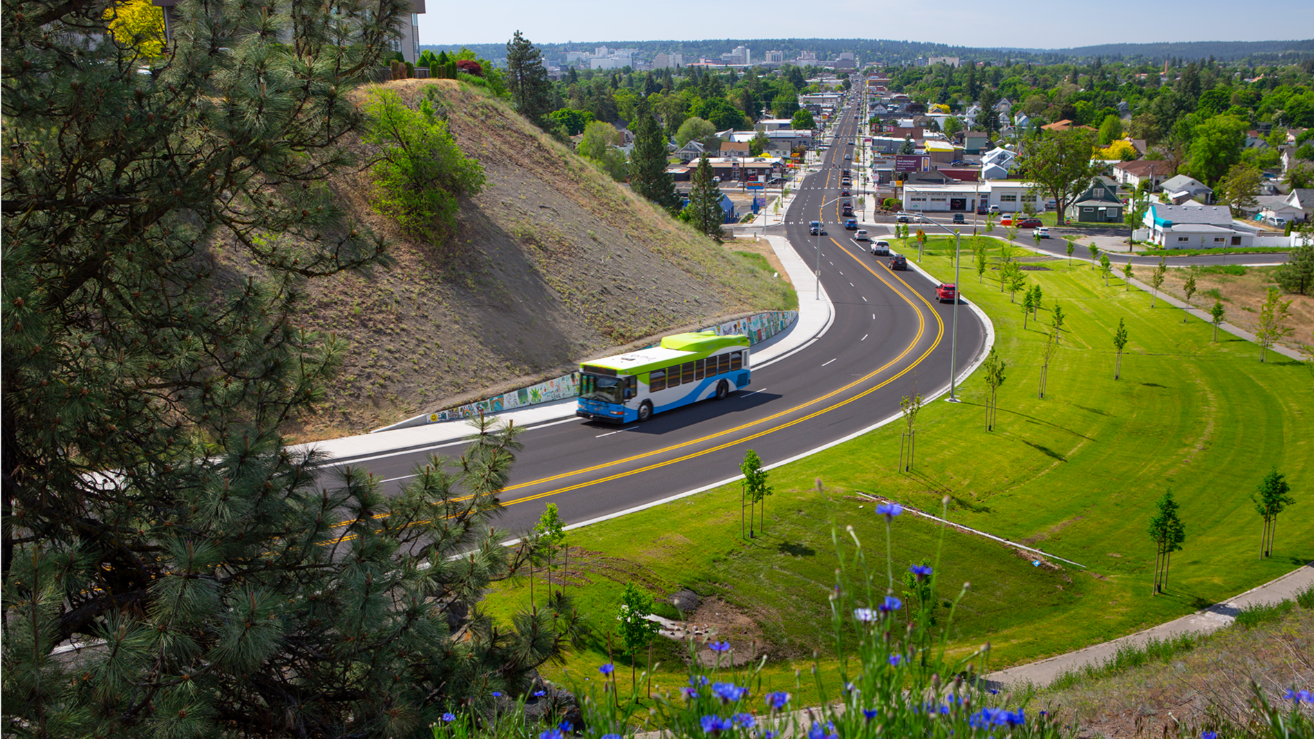 Aerial view of a city road with a Spokane Transit bus traveling, surrounded by lush greenery and a hill, with houses and trees in the background under a clear sky.