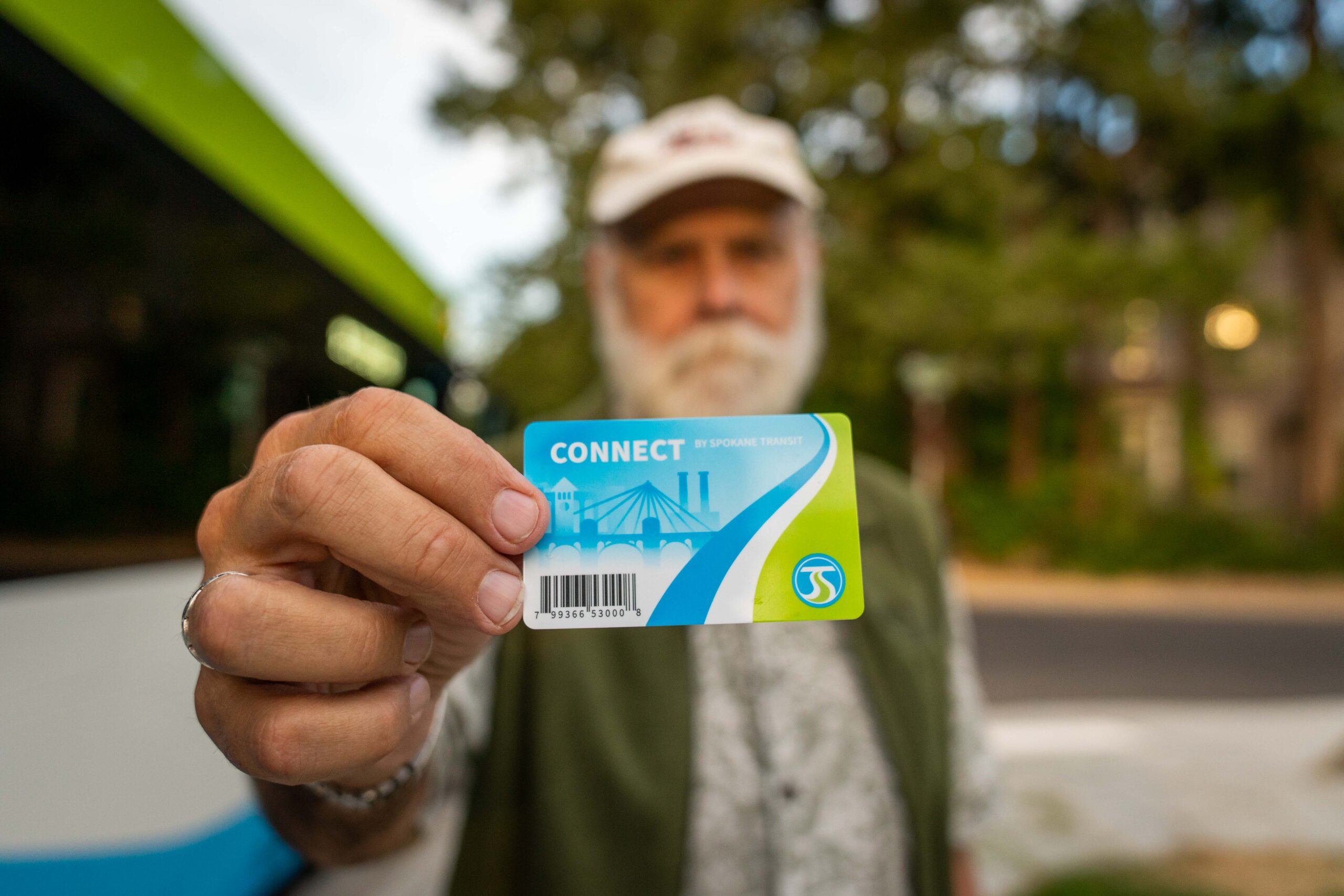 A bearded man wearing a cap holds a "CONNECT" public transit card towards the camera, displaying it prominently. The background shows blurred greenery and a bus, suggesting he is at a bus stop or in a transit area.